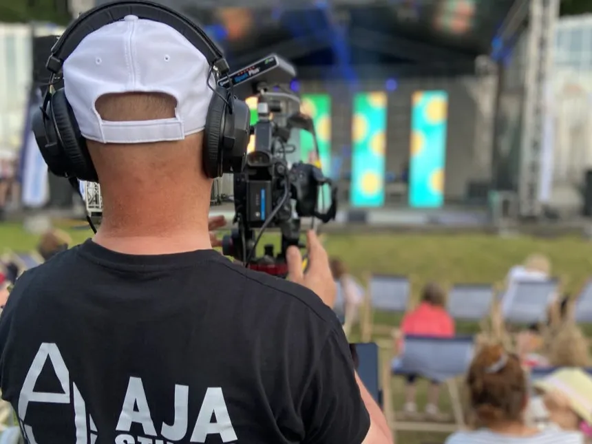 Operator w koszulce AJA Studio ze słuchawkami przy kamerze podczas wydarzenia