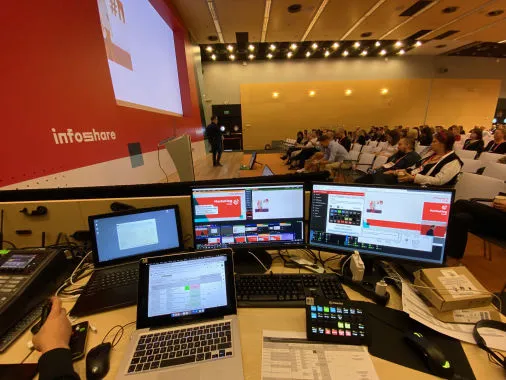 Stanowisko realizacyjne z monitorami i laptopami na sali konferencyjnej, w tle uczestnicy i ekran