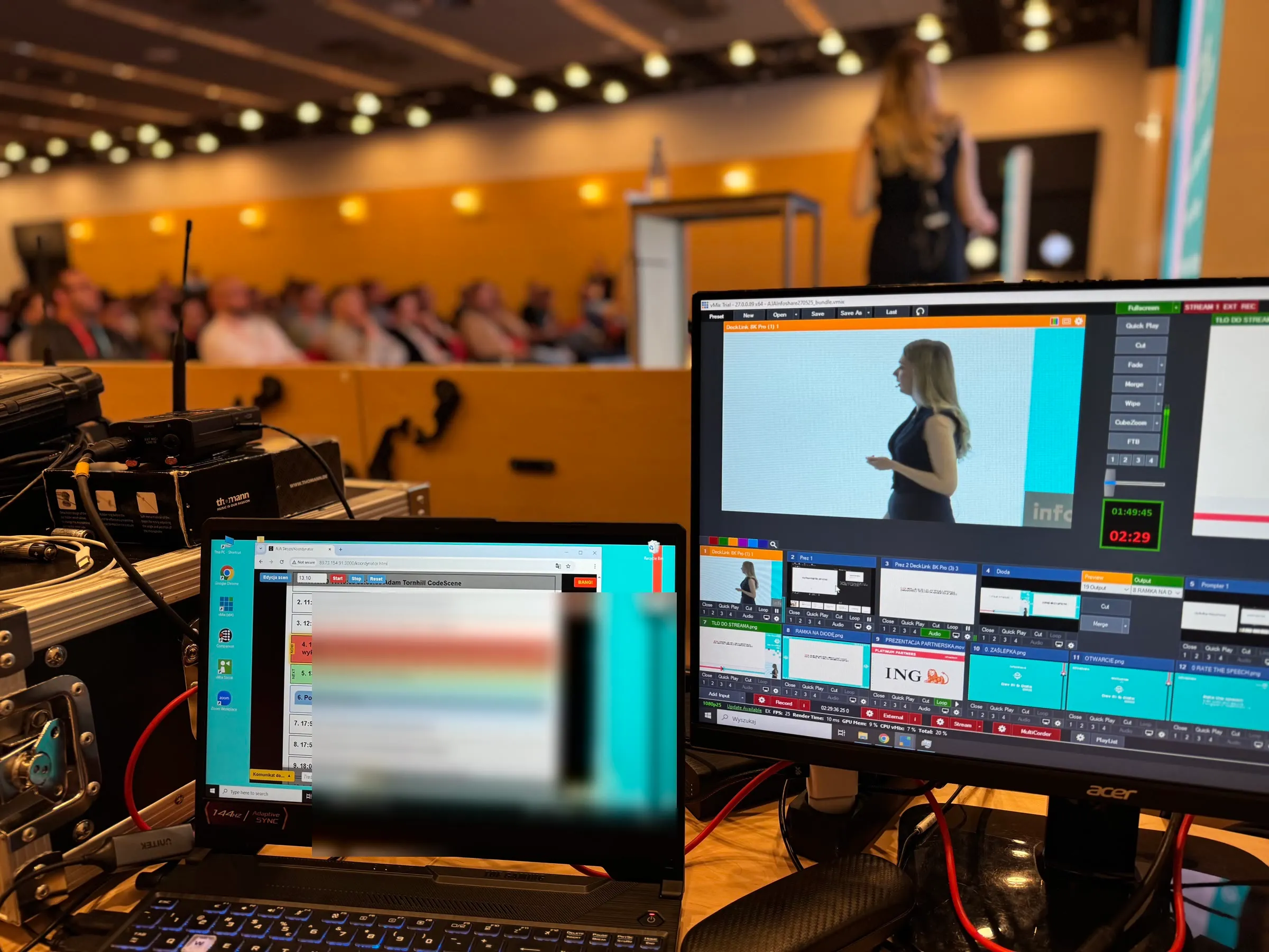 Stanowisko realizacyjne z laptopem i monitorem, w tle scena oraz publiczność konferencji