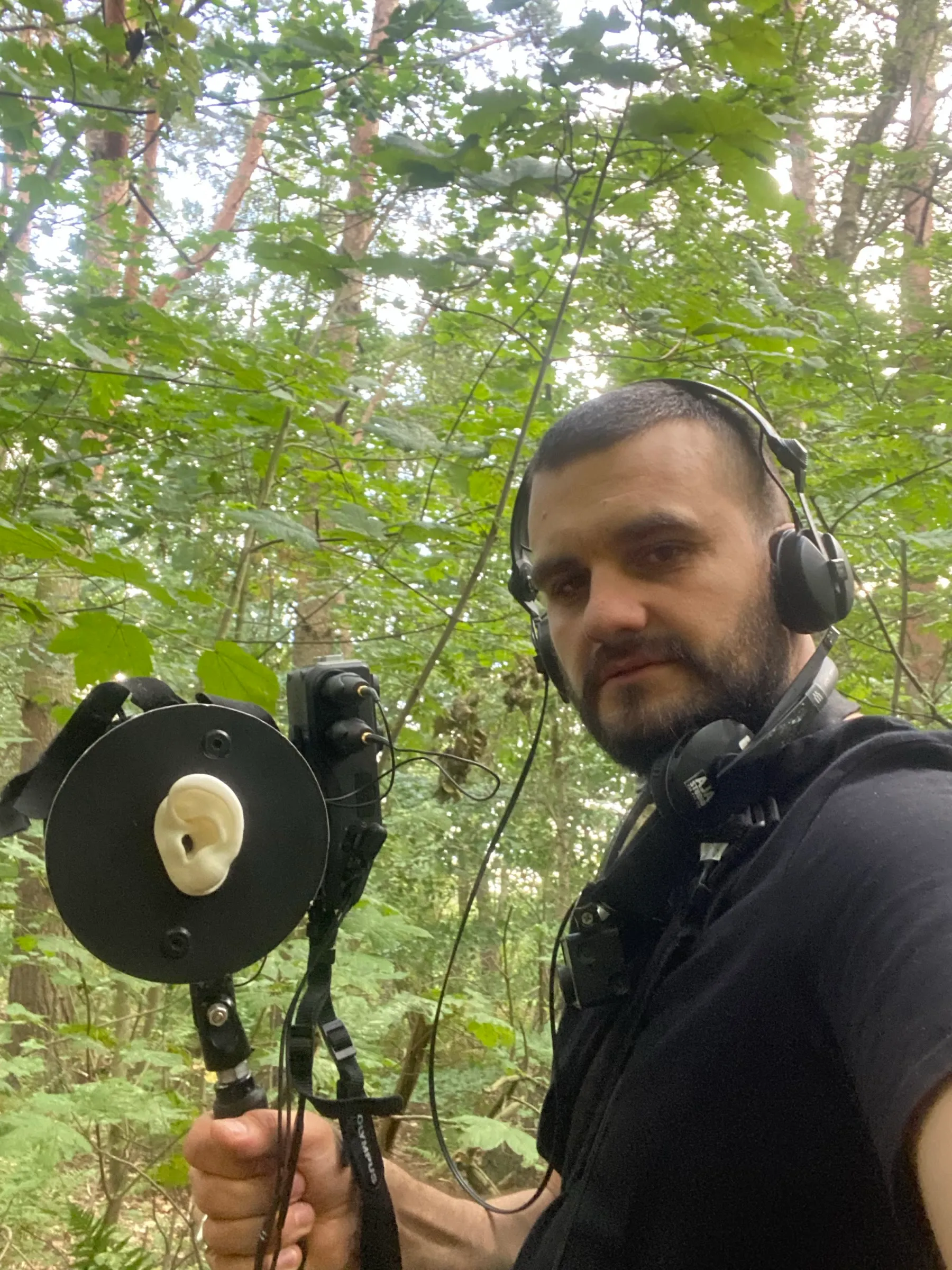 Adrian Jerzy Amrugiewicz w słuchawkach w lesie z mikrofonem binauralnym na statywie ręcznym