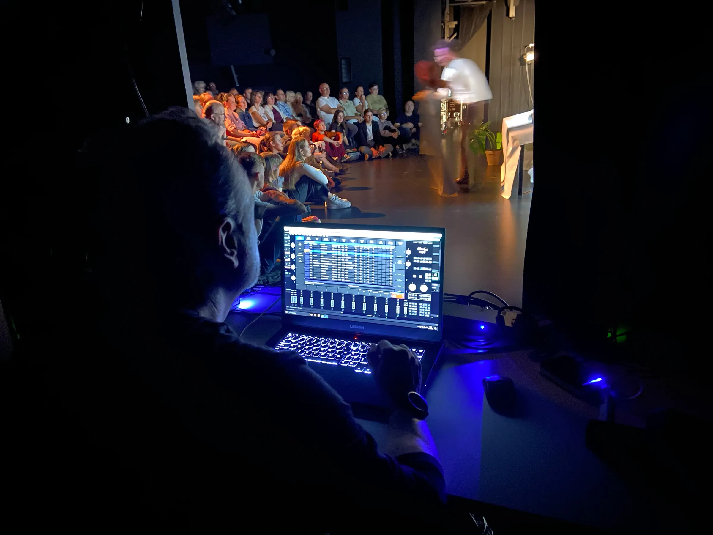 Operator przy laptopie z programem do obsługi dźwięku, za nim scena teatralna i publiczność