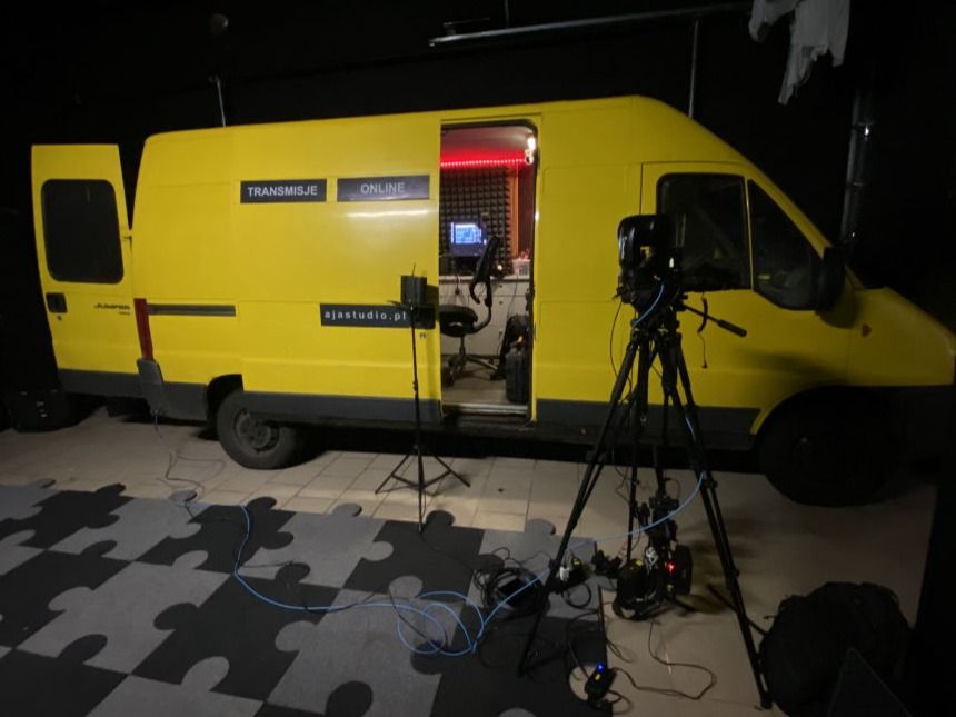 Żółty wóz realizacyjny AJA Studio z otwartymi drzwiami, obok kamera na statywie