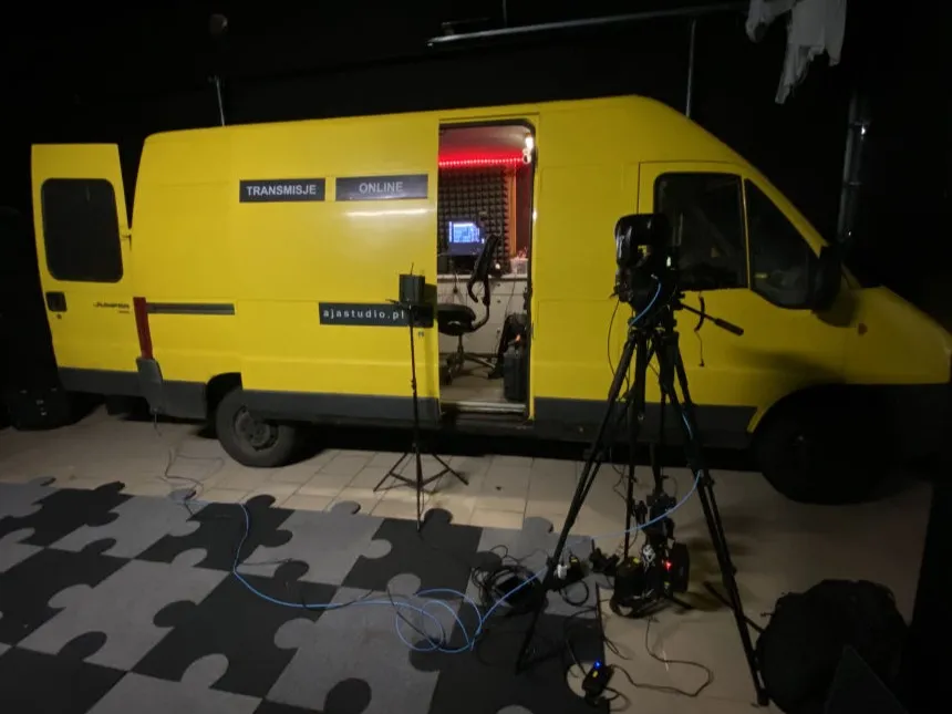 Żółty wóz realizacyjny AJA Studio z otwartymi drzwiami, obok kamera na statywie